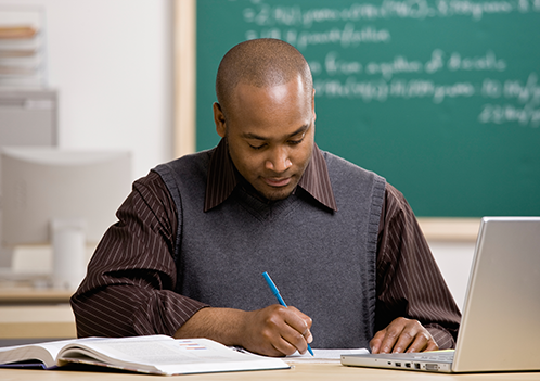 Instructor grading papers with laptop and paper and pen.